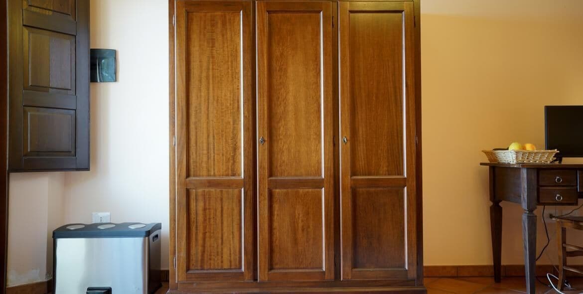 A room with a wooden wardrobe, a small trash bin, and a television on a stand.