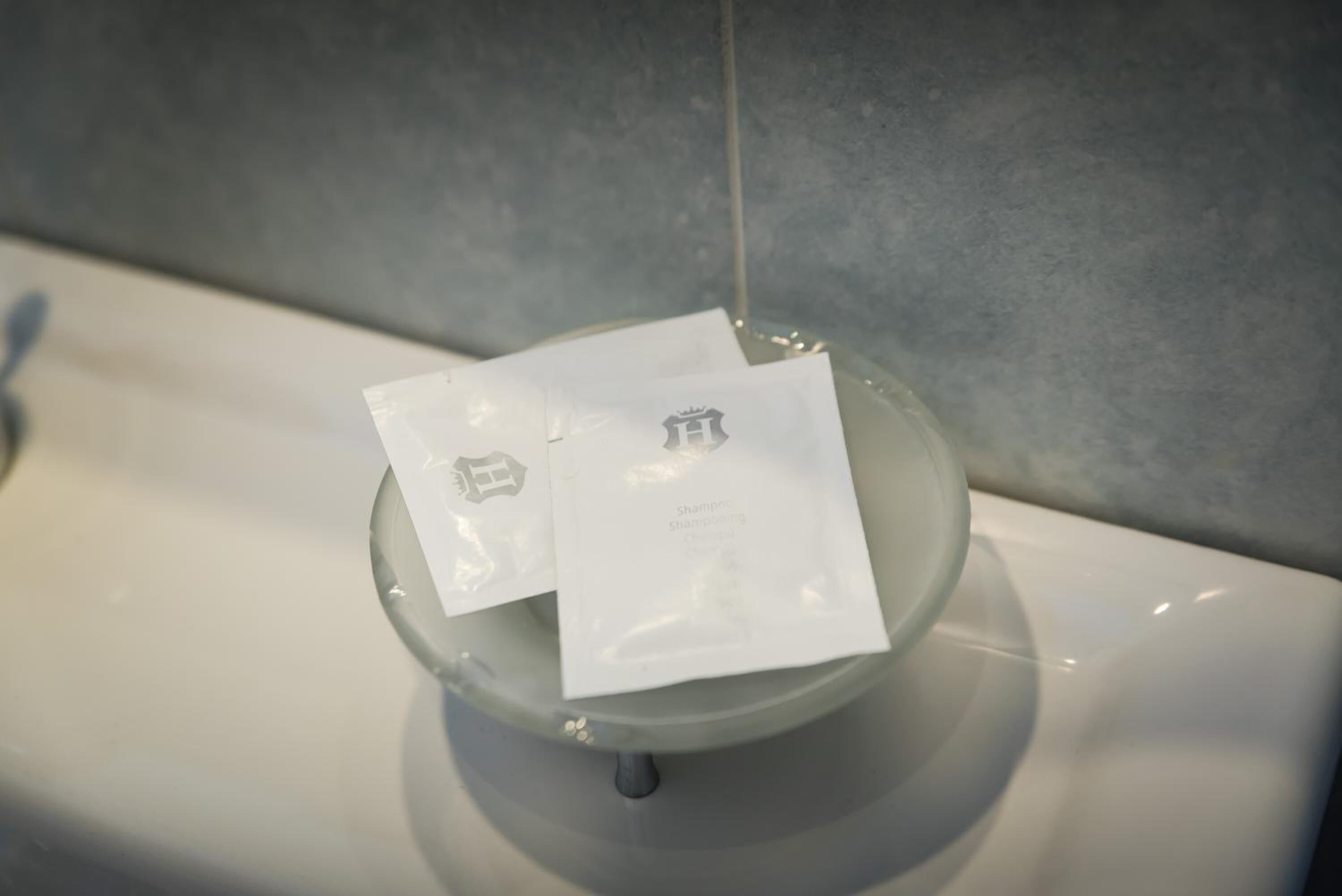 A glass bowl on a sink with two white packets labeled "Shampoo" and "Shampoo Conditioner".