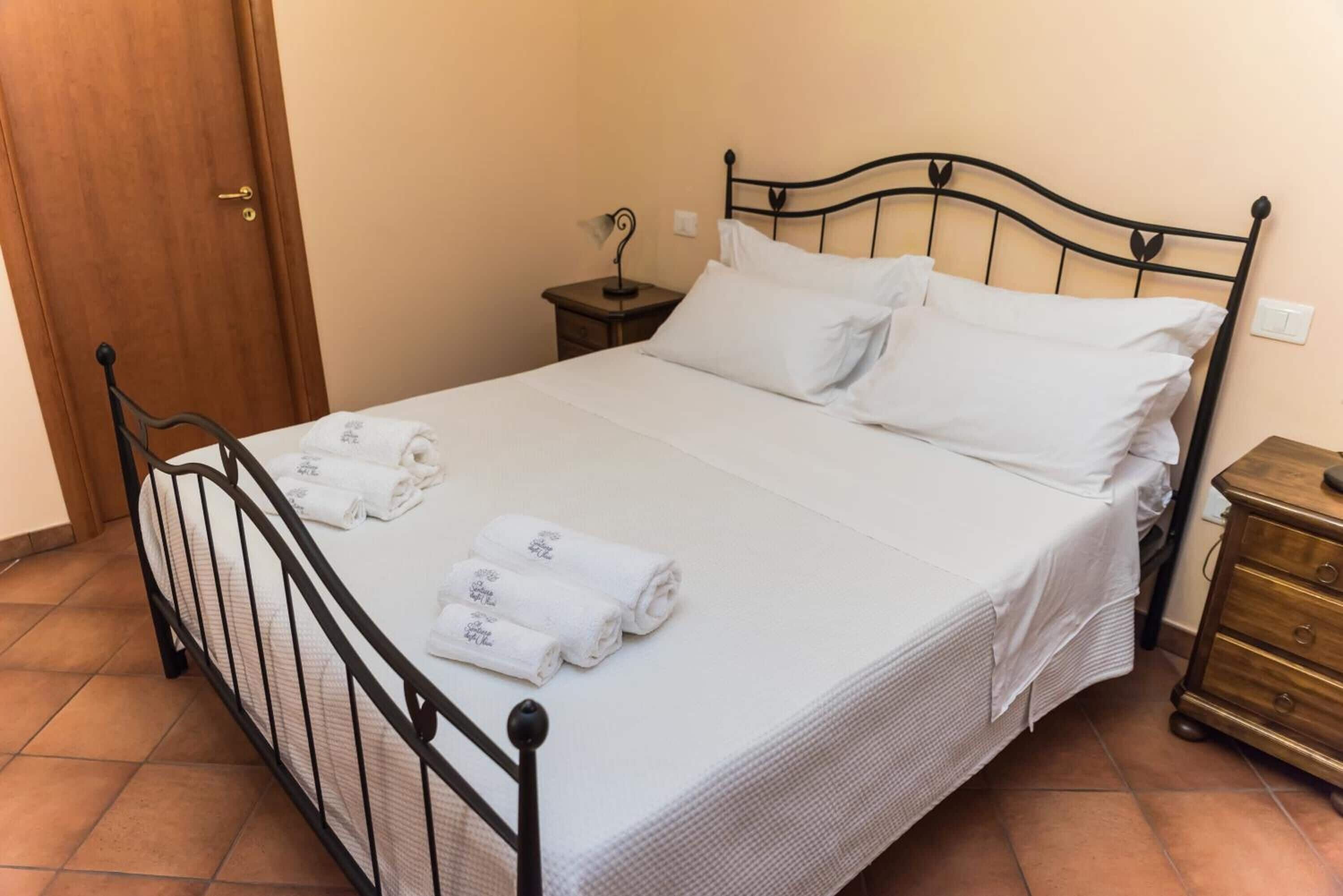 A neatly made bed with white linens and rolled towels, a wooden door, a small bedside table with a lamp, and a wooden dresser.
