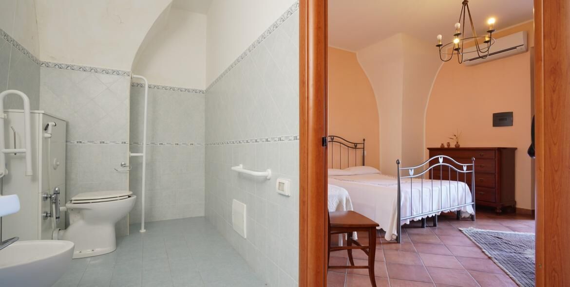 A bathroom with a toilet, shower, and sink. The room has a bed with a wooden headboard and a bedside table.