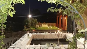 An outdoor dining area with tables set for a meal, surrounded by greenery and a building in the background.