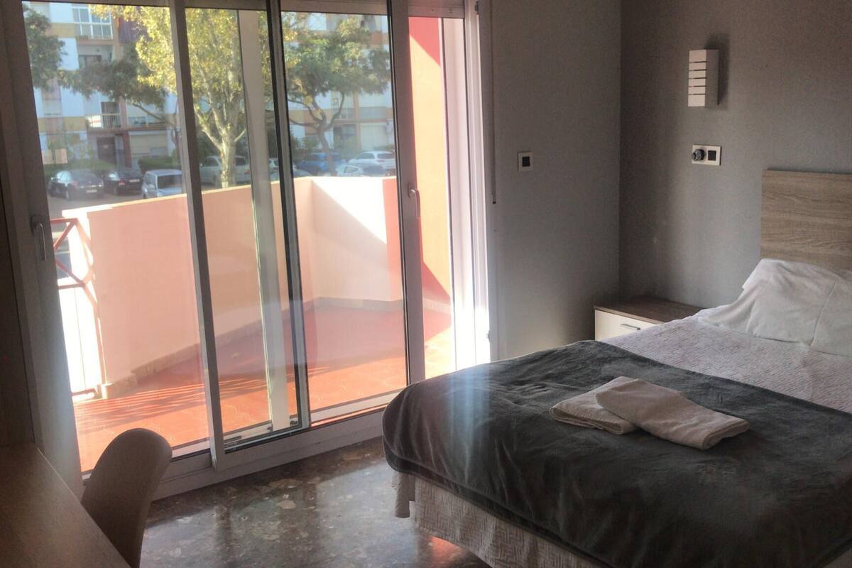 Habitación de hotel con cama, mesita de noche, una silla y puerta corrediza de vidrio que da a un espacio exterior.