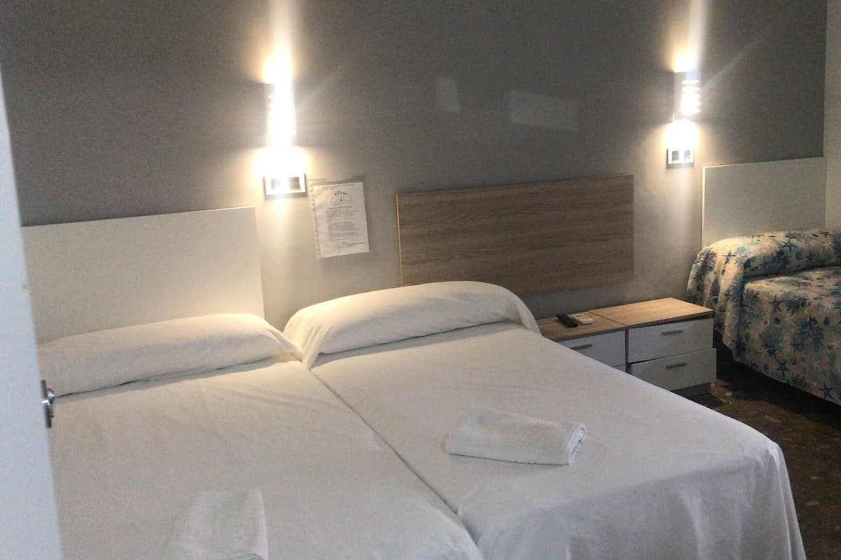 Habitación de hotel con dos camas individuales, cabecero de madera, mesitas de noche con lámparas y una pequeña mesita auxiliar.