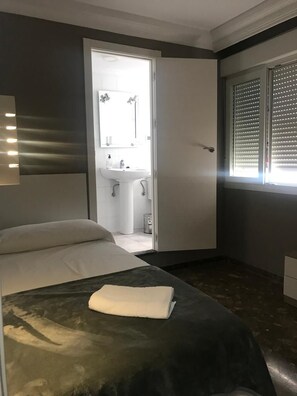 Une chambre d’hôtel avec un lit, une salle de bain comprenant un lavabo et un miroir, et une fenêtre avec des stores.