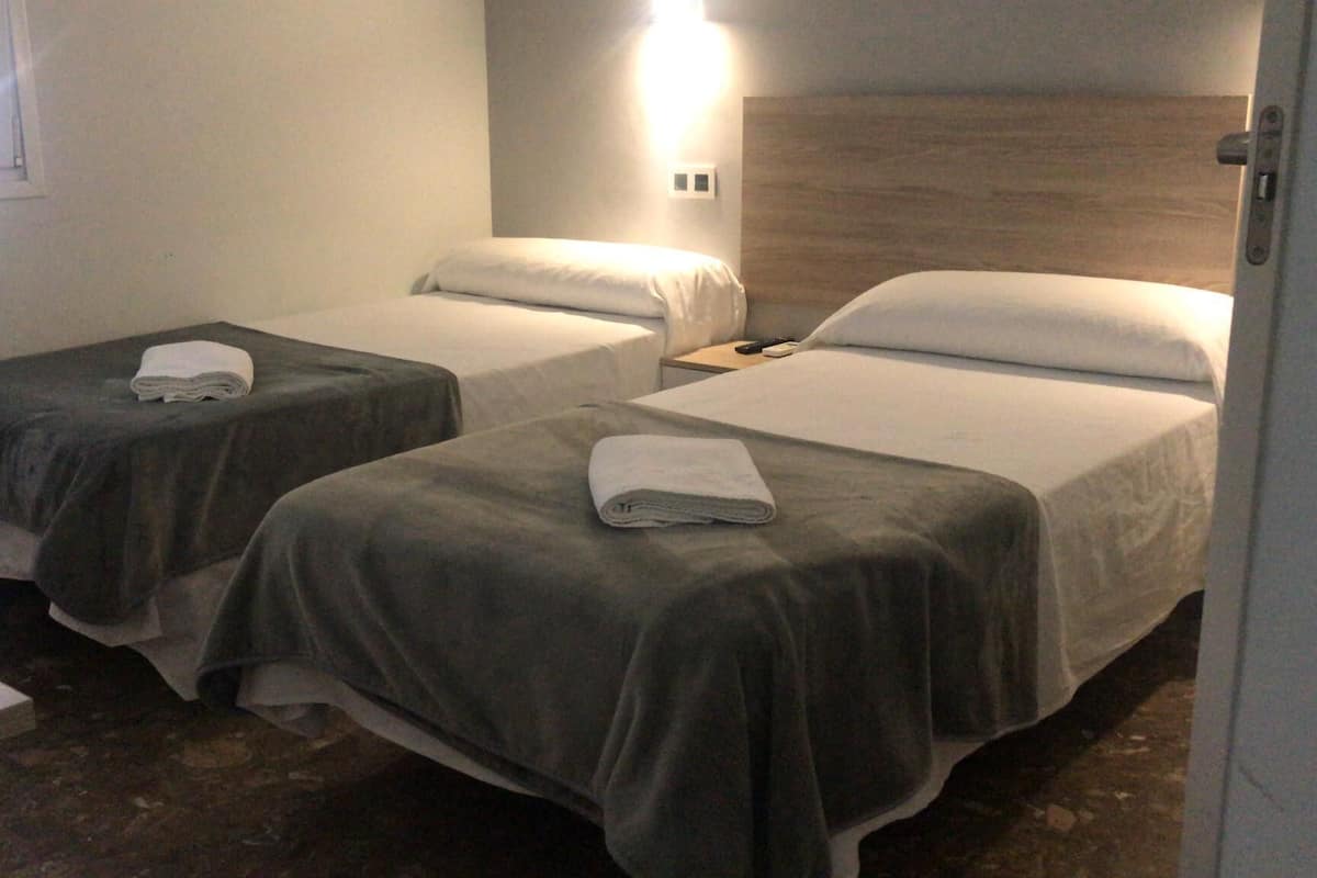 Habitación de hotel con dos camas individuales, cada una con una manta gris y un almohadón blanco. Se observa una mesita de noche con una lámpara y un teléfono.