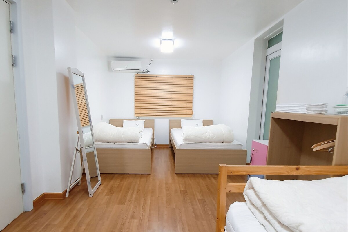 A room with two beds, a wooden floor, a mirror, and a window with blinds.