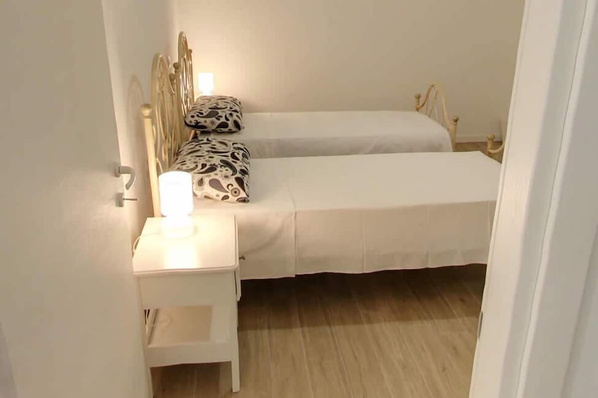 Un dormitorio pequeño y ordenado con dos camas individuales, una mesita de noche con lámpara y una silla.