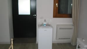 Een kleine kamer met een witte koelkast, een raam met zonwering, een radiator en een deur met een handvat.