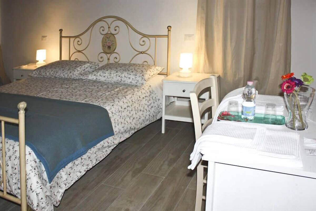 Un dormitorio con cama, mesitas de noche, una silla y una mesa con un florero.