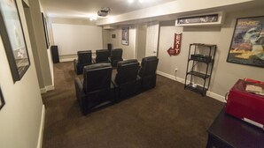 A home theater room with a projector, multiple black reclining chairs, a red storage box, and a poster on the wall. - Ski Home with Hot Tub and King Beds (Sandy)