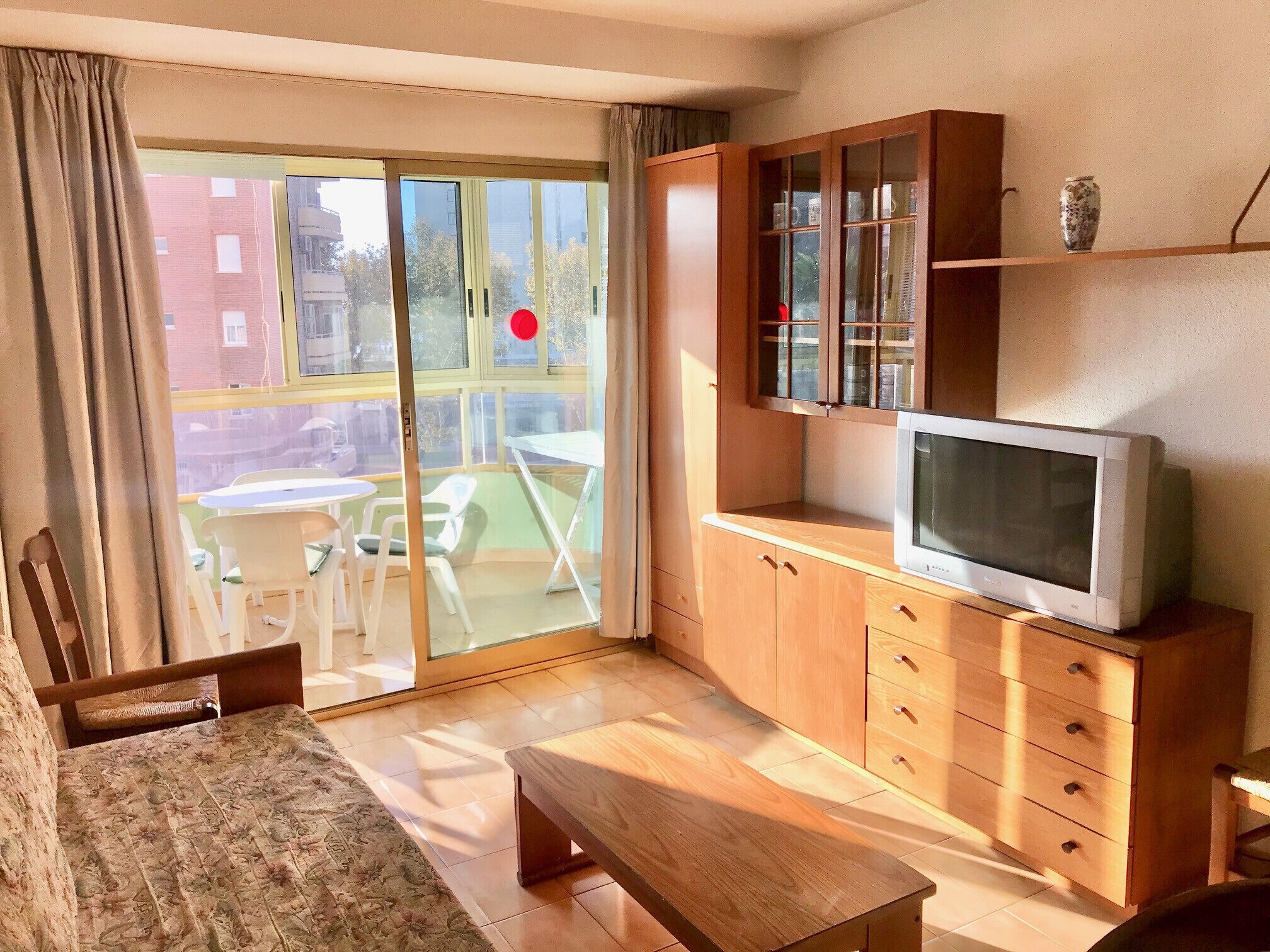 A living room with a wooden cabinet, a television, a bench, and a sliding glass door leading to an outdoor area with chairs and a table.