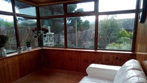Ein Eckzimmer mit einer weißen Ledersofa, Holzwandverkleidung, einem großen Fenster mit Blick ins Freie und einem kleinen Tisch mit Blumen in einer Vase.