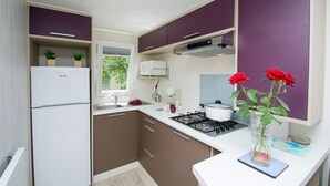 A compact kitchen with a refrigerator, stove, microwave, and sink. There are cabinets, a window, and a vase with flowers on the counter. - Camping Le Vieux Colombier (Moustiers-Sainte-Marie)