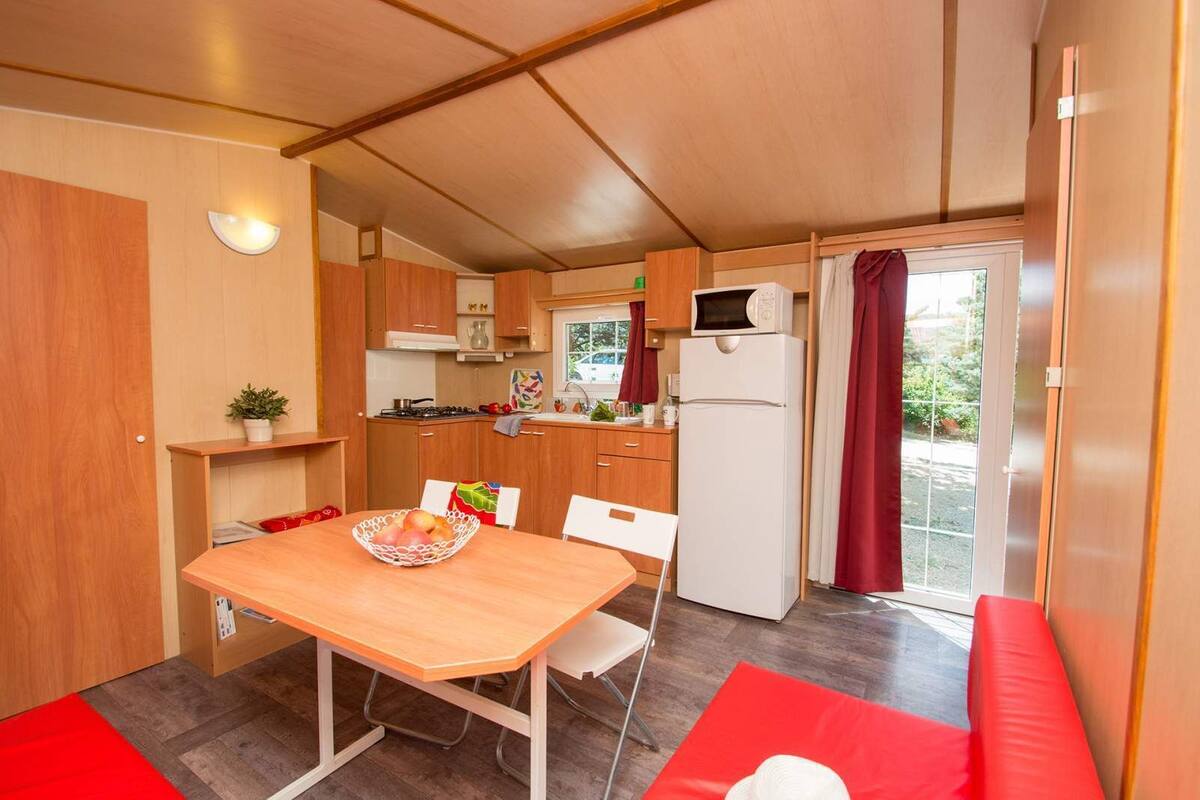 A compact kitchenette with a microwave, stove, and sink. There is a dining table with chairs and a red cushion.