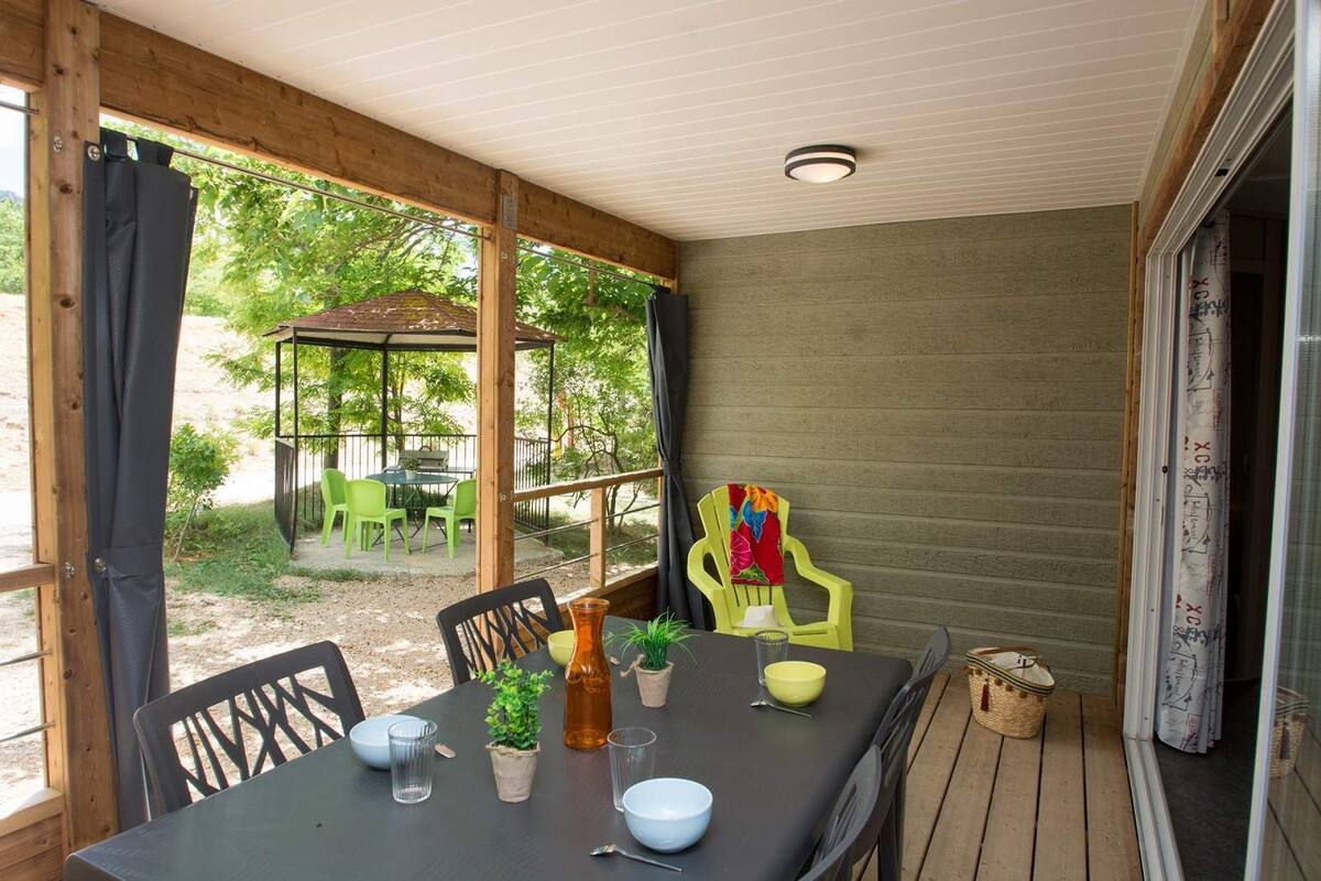 A covered outdoor dining area with a table set for two, chairs, and a small garden visible through the open sides.