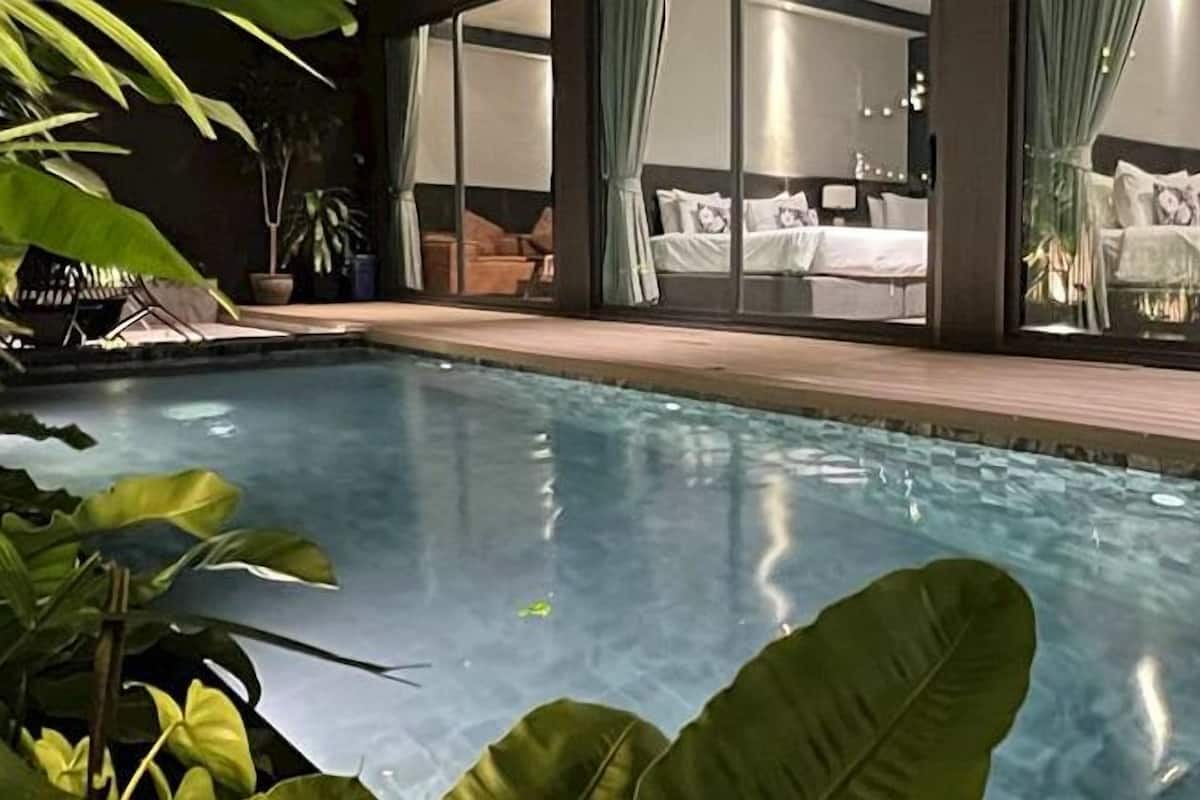 A swimming pool with a wooden deck, surrounded by large green plants.