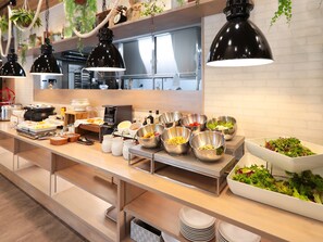 A buffet setup with various food items and utensils, including bowls of salad and other dishes, arranged on a long table with hanging pendant lights. - Green Rich Hotel Yonago Ekimae Artificial Hot Spring Futamata Yunohana (Yonago)