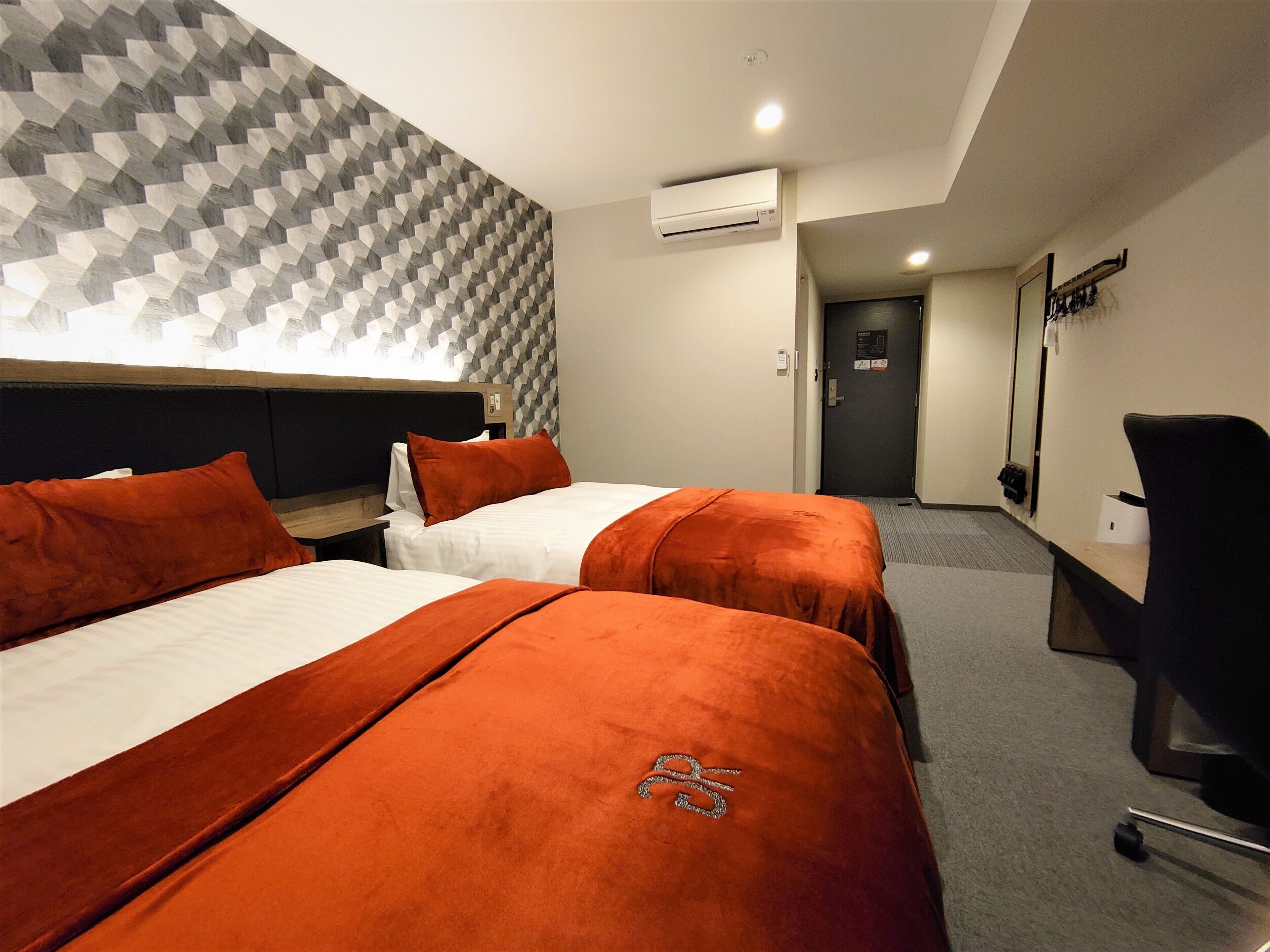 Ein Hotelzimmer mit zwei Betten, jeweils mit einer orangefarbenen Tagesdecke und passenden Kissen. An der Wand befindet sich ein Klimagerät und eine Tür zu einem Flur. Ein geometrisches Muster ziert den Kopfteil.