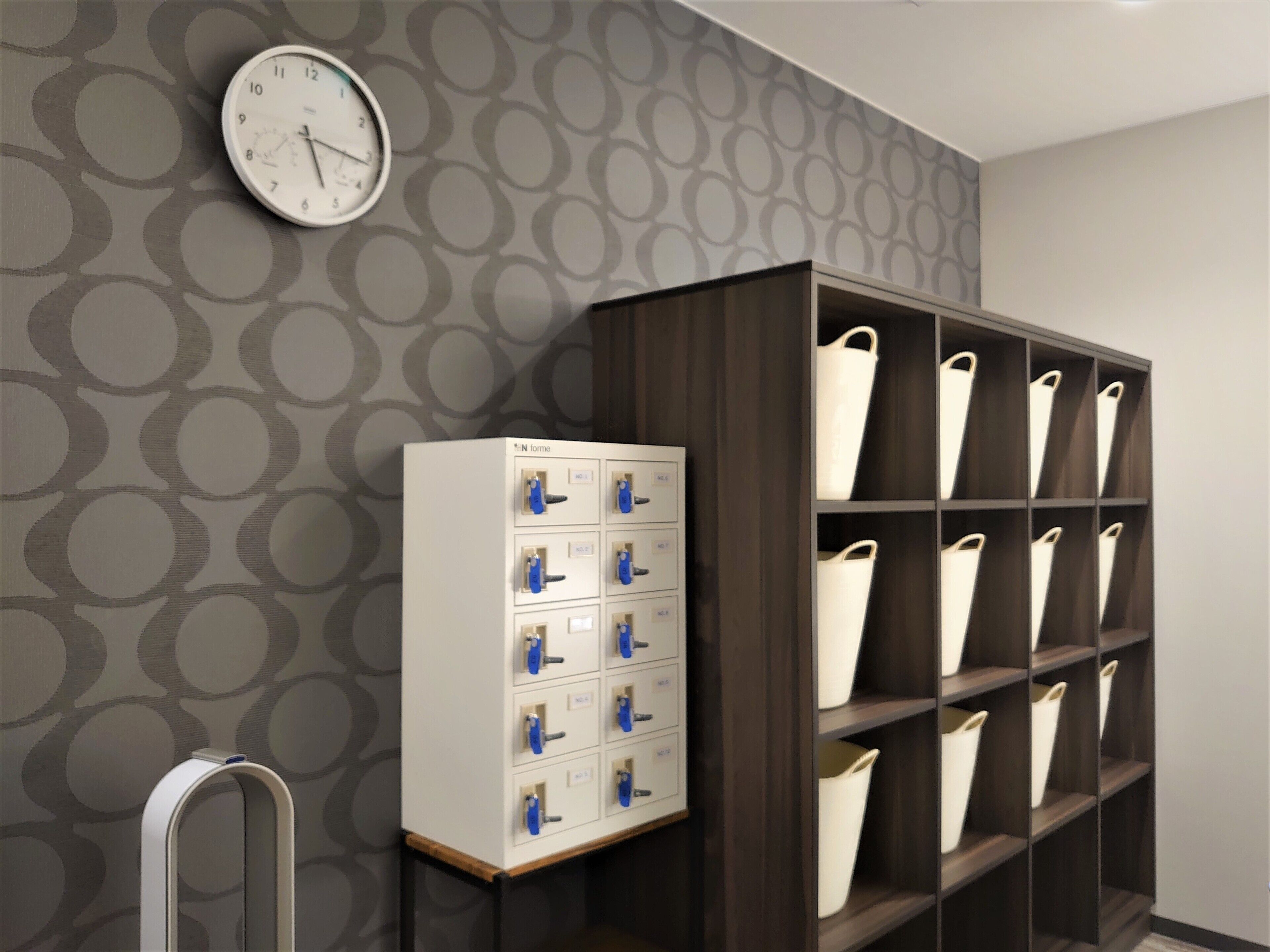 A room with a patterned wallpaper, a clock, a metal cabinet with drawers, and a shelving unit with baskets.