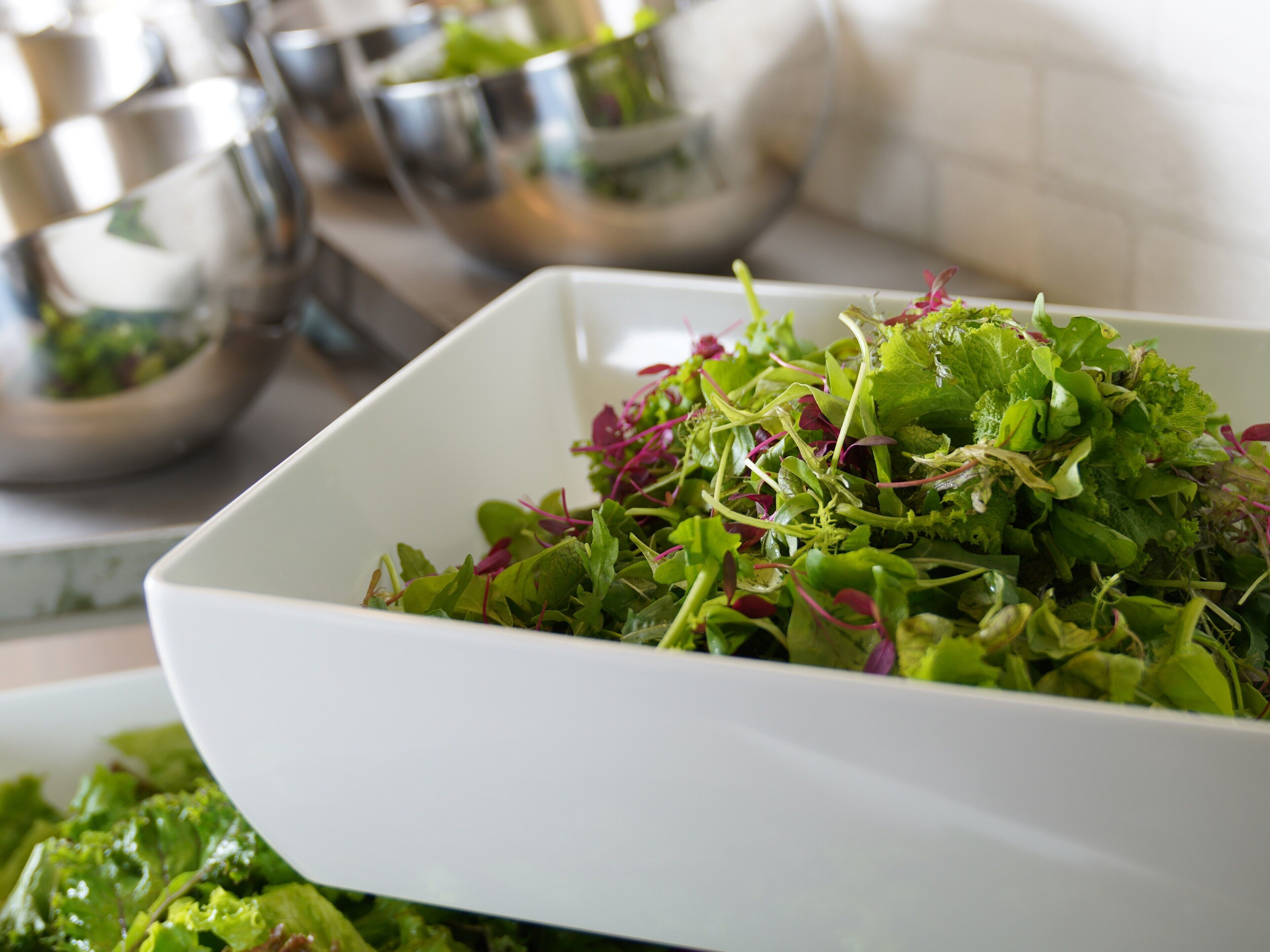 A white bowl filled with mixed green and red leafy vegetables, placed on a kitchen counter with other bowls in the background.