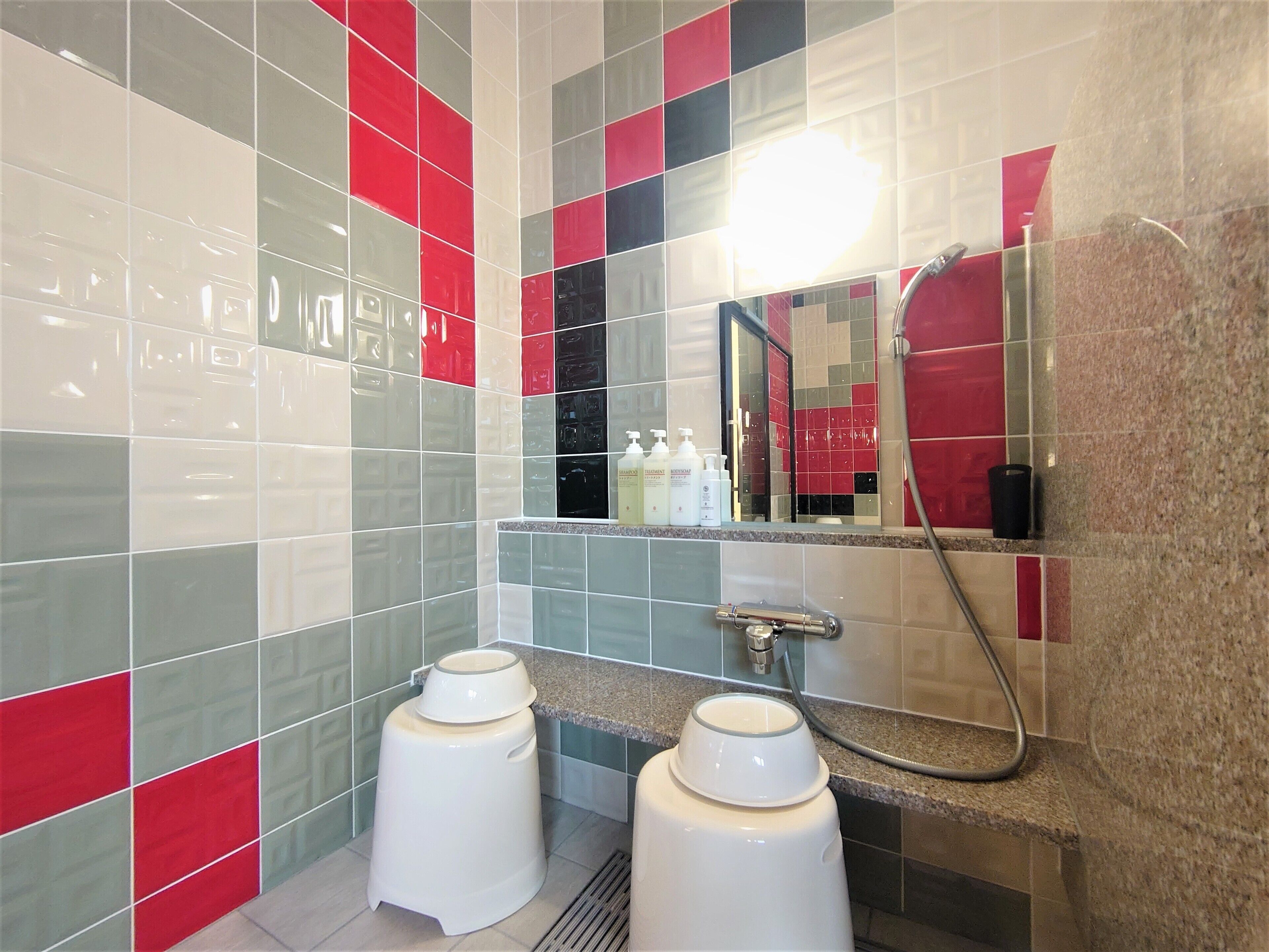 A bathroom with tiled walls in red, grey, and white, a granite countertop, two white pedestal sinks, and a handheld showerhead.