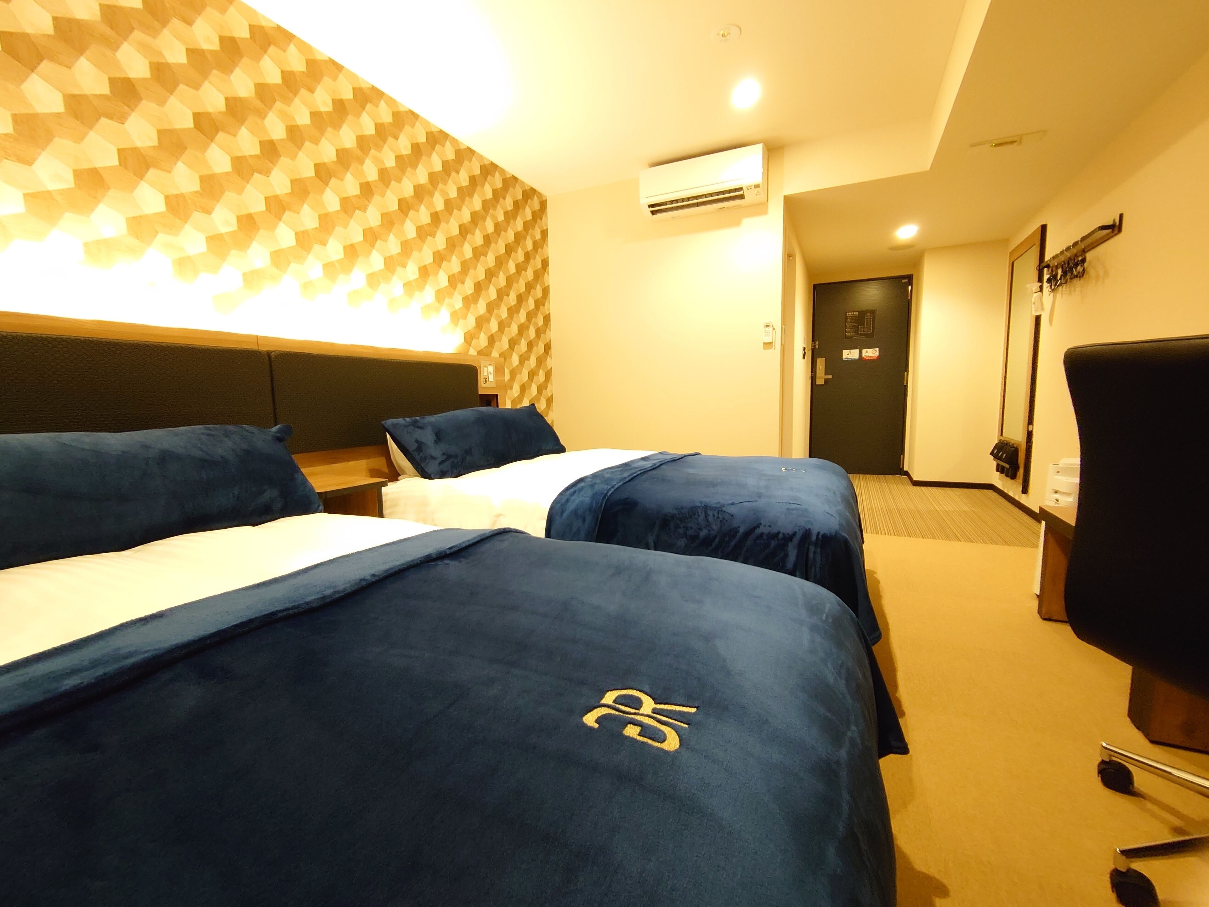 A hotel room with two beds, a desk, a chair, and a patterned wall.