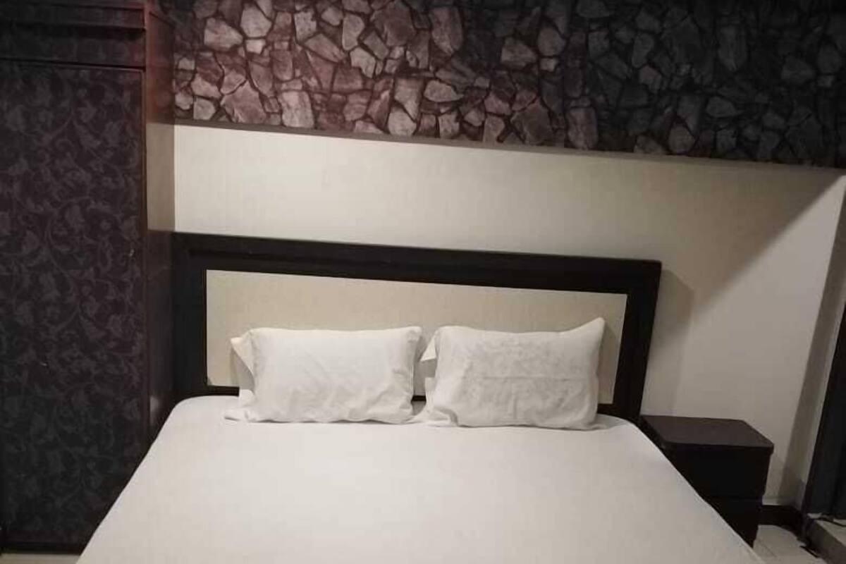 A bed with a white mattress and two pillows, a dark headboard, and a stone-patterned wall above.