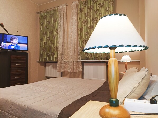 A hotel room with a bed, a wooden nightstand with a lamp, a television on a dresser, and patterned curtains.
