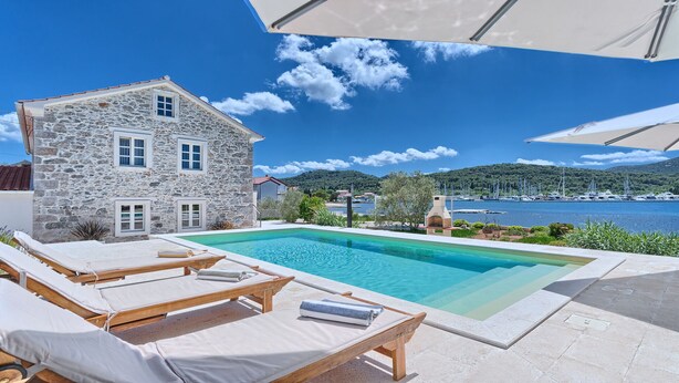 Une maison en pierre avec une piscine, des chaises longues et une vue sur les bateaux qui naviguent.