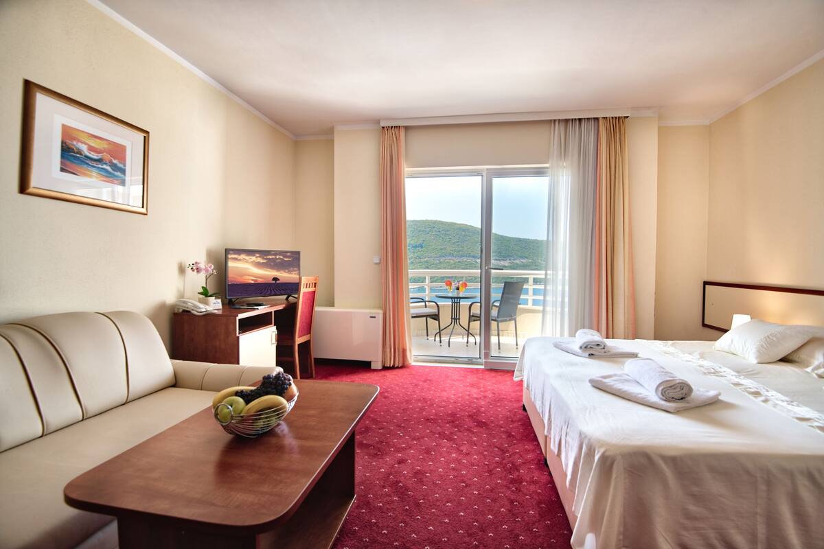 A hotel room with a bed, a sofa, a desk with a computer, a small table with fruit, and a balcony with a table and chairs.