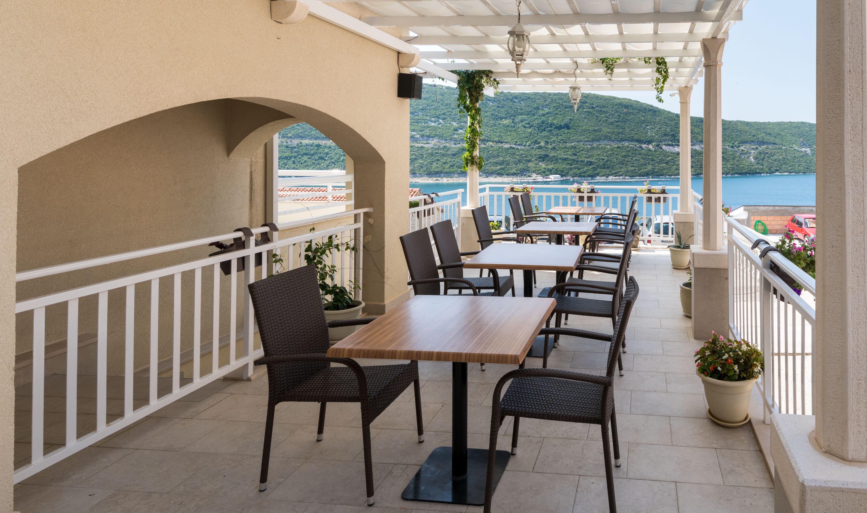 Una terraza al aire libre con mesas y sillas, una pérgola y vistas al mar y las colinas.