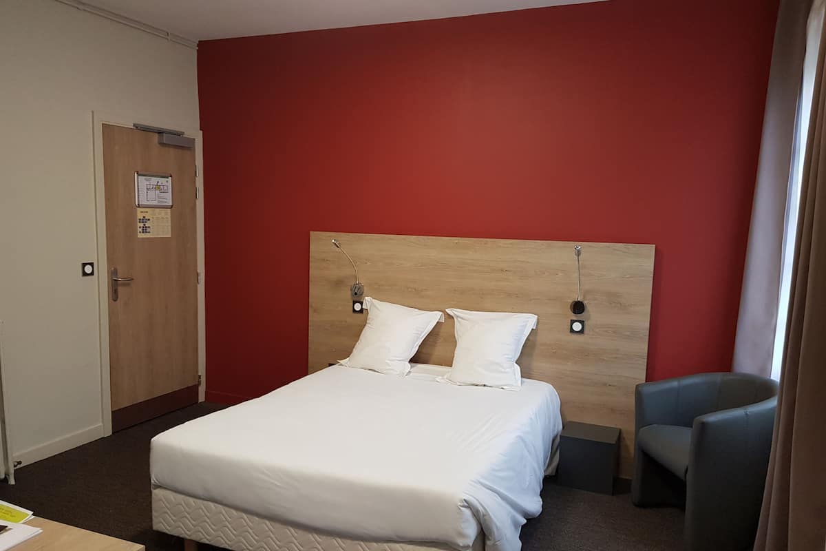 Habitación de hotel con cabecera de madera, dos almohadas blancas, un sillón gris y una pequeña mesa con papeles.