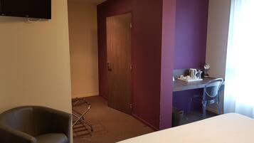 A hotel room with a bed, a desk with a chair, a TV mounted on the wall, and a door leading to another room.