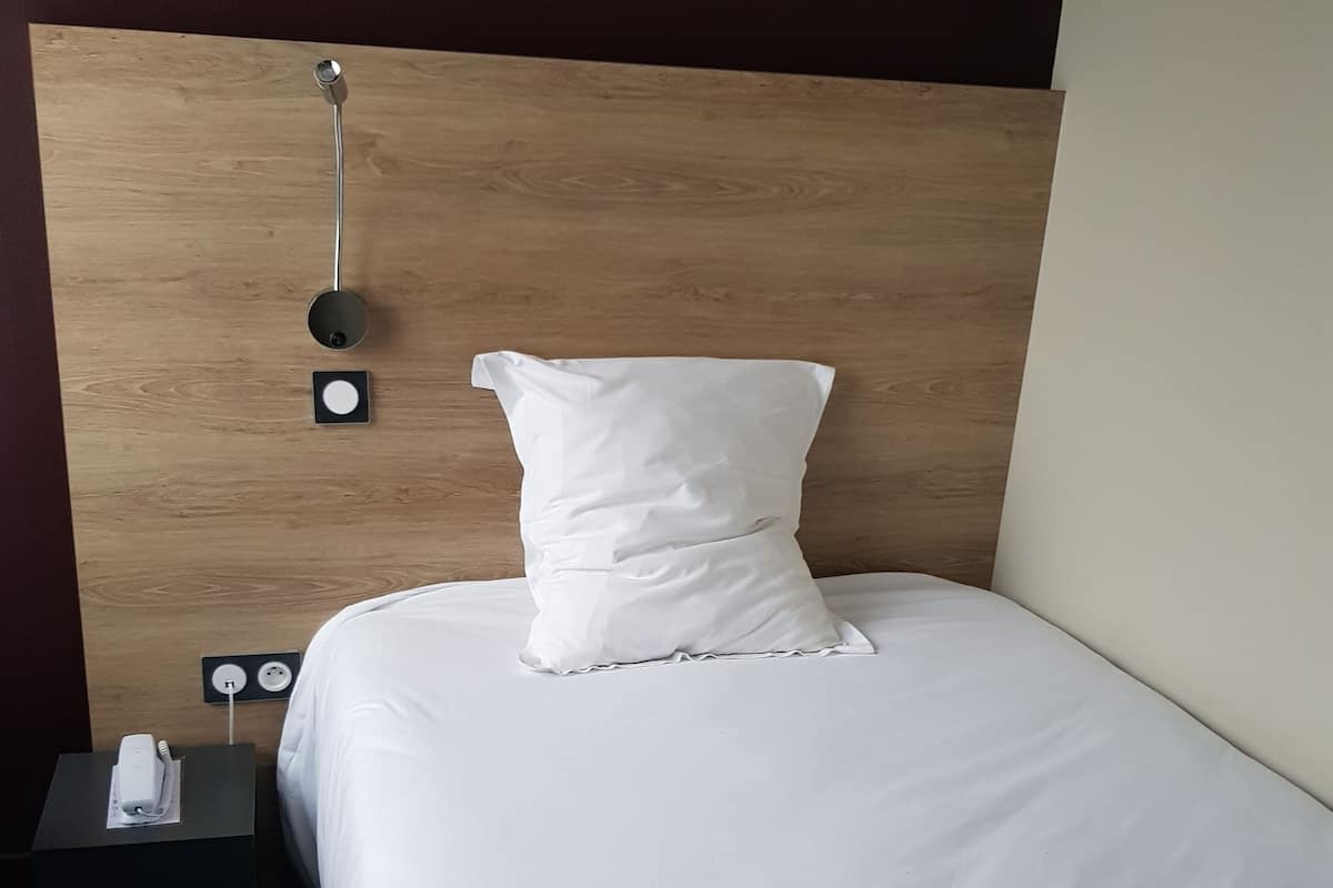 Una cama bien hecha con ropa blanca y almohadas, una mesita de noche con enchufe y teléfono, y una lámpara de pared.