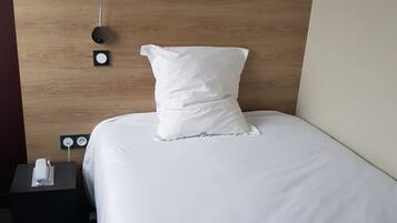 Una cama bien hecha con ropa blanca y almohadas, una mesita de noche con enchufe y teléfono, y una lámpara de pared.
