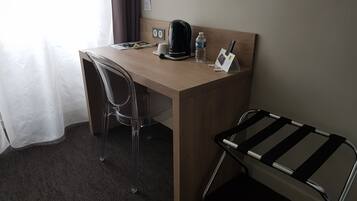 Una habitación de hotel con un escritorio de madera, una silla de plástico transparente, una silla de metal blanco y negro, una cafetera, una botella de agua y un control remoto.