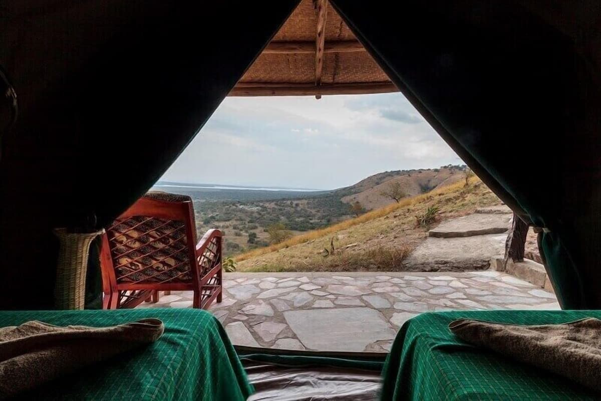 Quarto com vista para uma paisagem montanhosa, piso de pedra e duas camas com cobertores verdes.