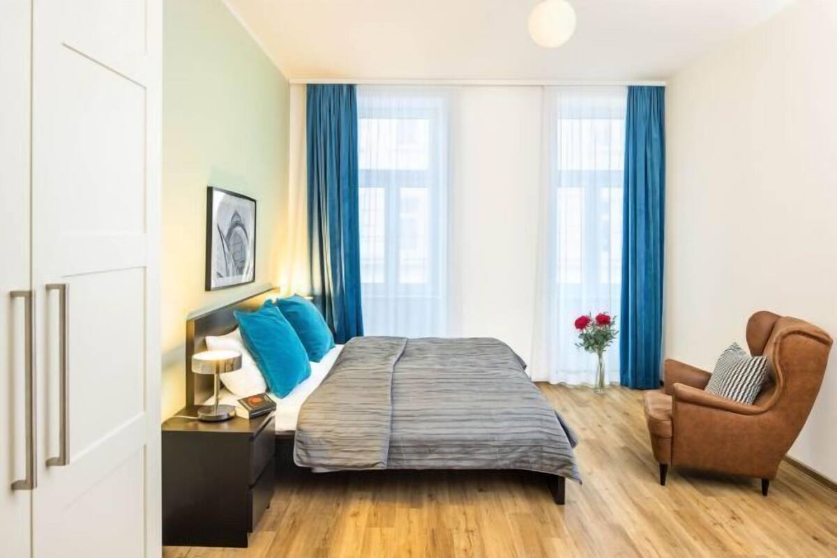 Une chambre à coucher avec un lit, une chaise, une table de chevet, une fenêtre avec des rideaux et un tableau encadré au mur.