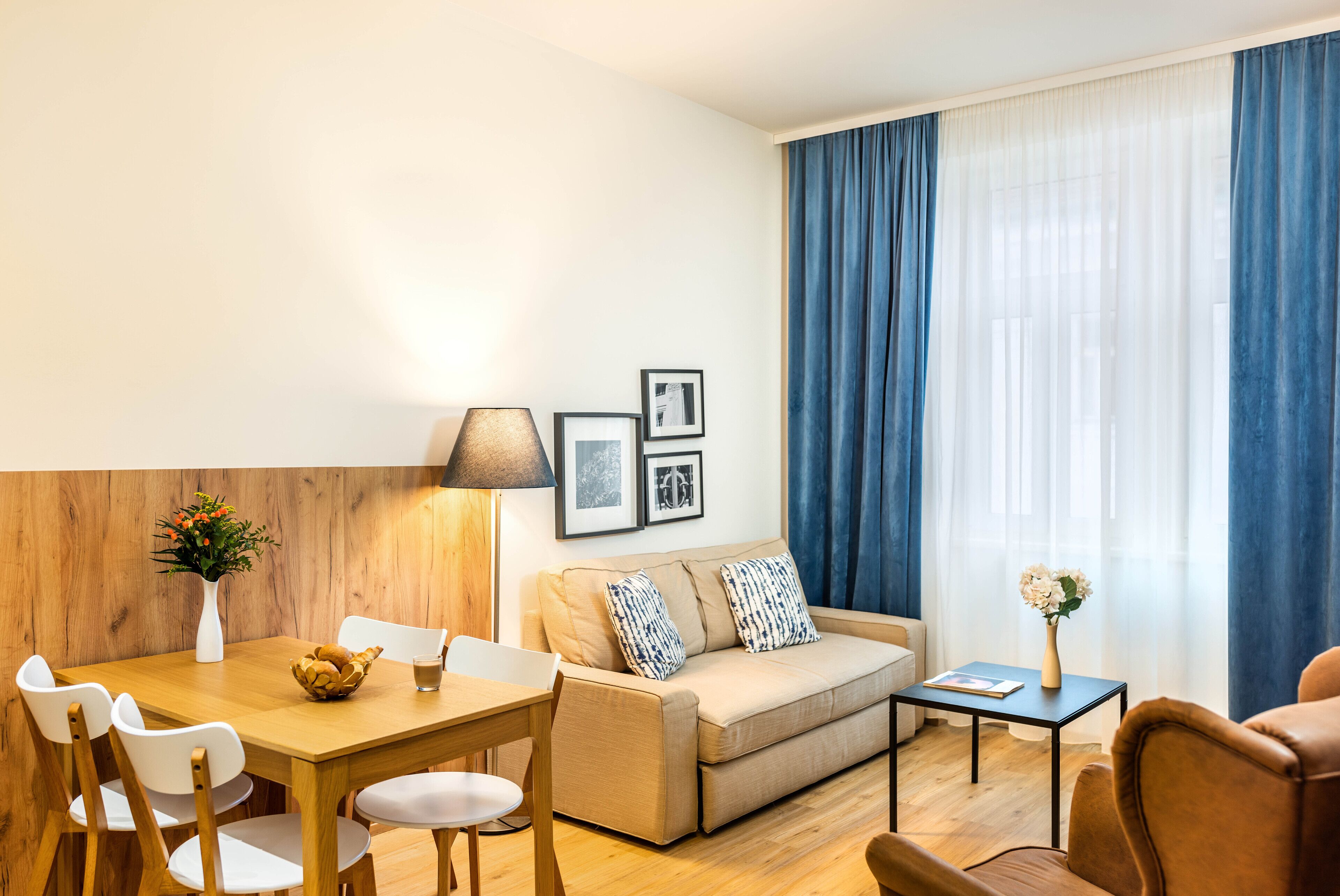 A modern living room with a wooden dining table, a sofa, and a coffee table. There are framed pictures on the wall and a lamp beside the sofa.