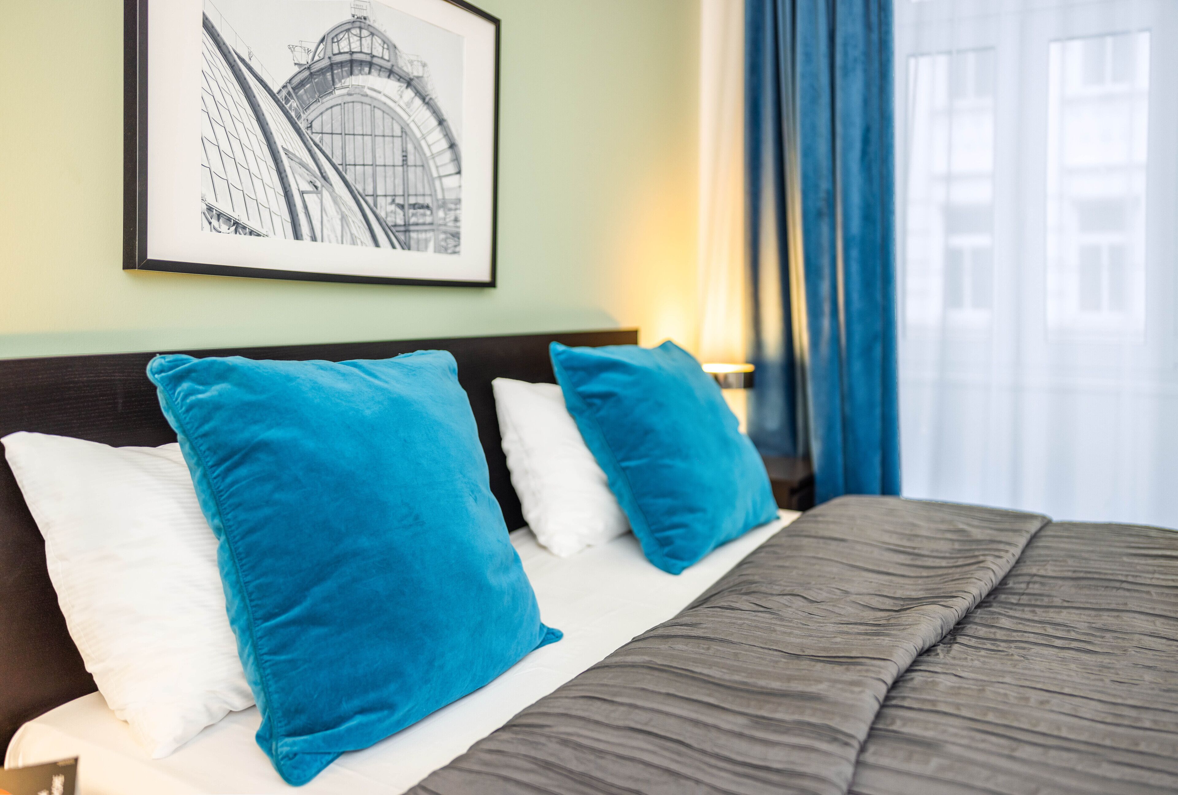 A neatly made bed with a dark headboard, two blue pillows, and a white pillow. A framed picture hangs on the wall above the bed.
