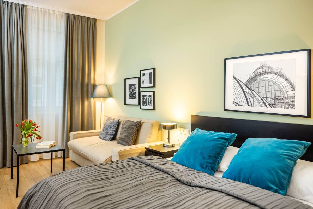 Une chambre d’hôtel avec un lit, une table de chevet avec une lampe, une petite table avec un vase de fleurs et un mur orné de tableaux.