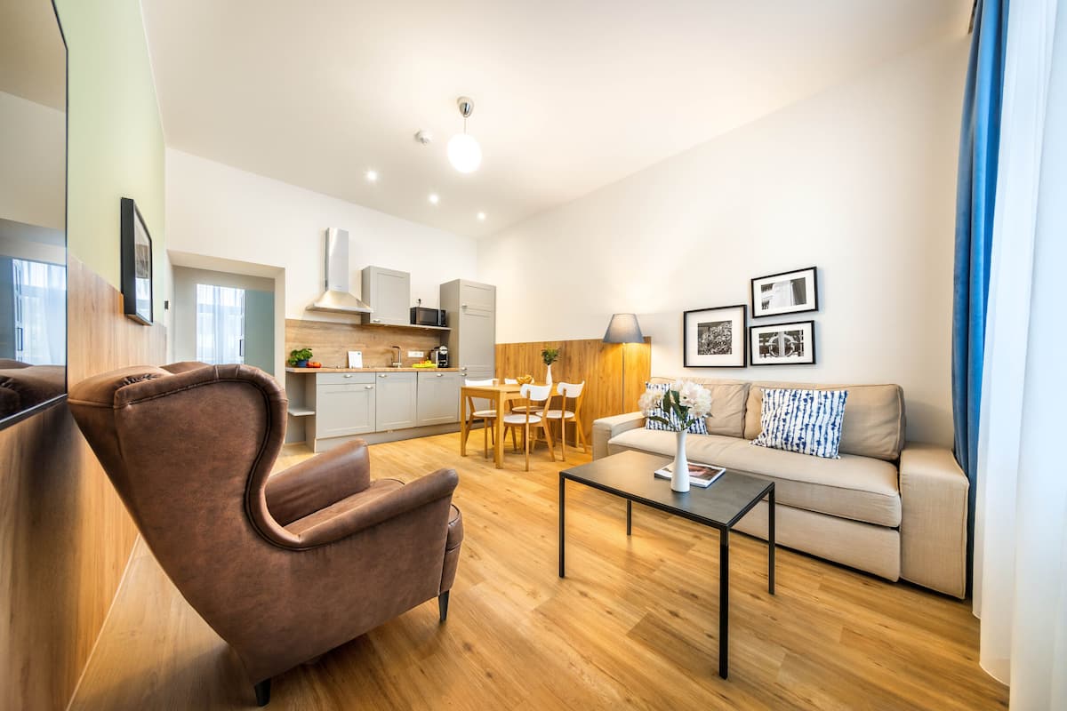 Un salon moderne avec un canapé, un fauteuil et une table basse. On y trouve également un coin repas avec une table et des chaises, et une cuisine à l’arrière-plan.