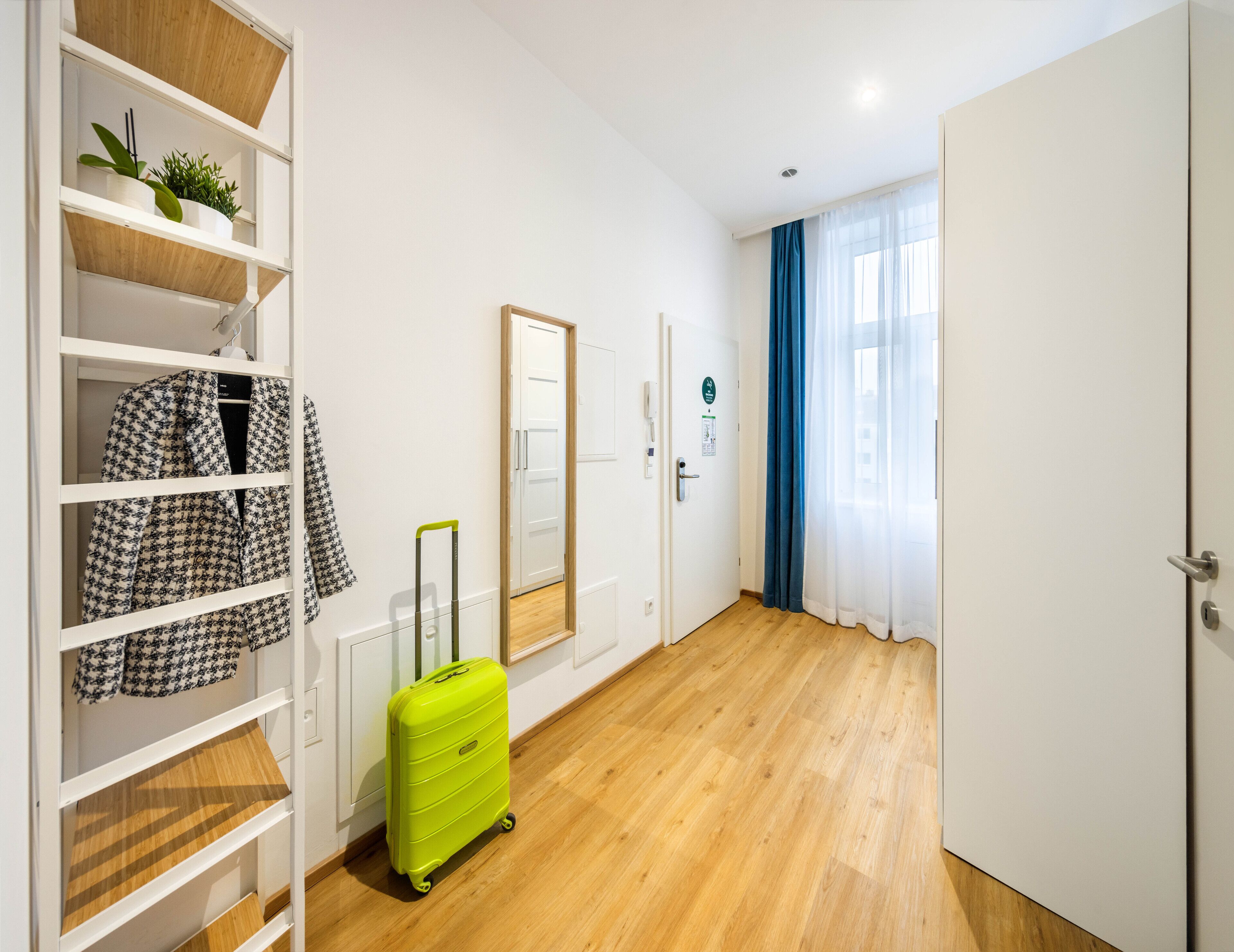 A hotel room with a wooden floor, a green suitcase, a white wardrobe with shelves, and a door with a blue curtain.