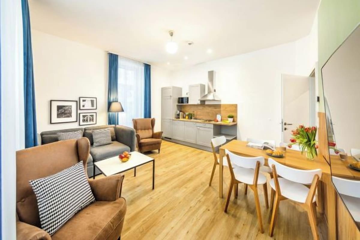 A modern living room with a sofa, armchair, and dining table. There is a kitchen in the background with cabinets and a stove.