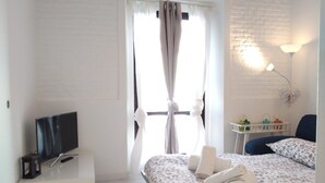 Quarto com cama, televisão sobre uma cômoda, janela com cortinas e luminária fixada na parede.