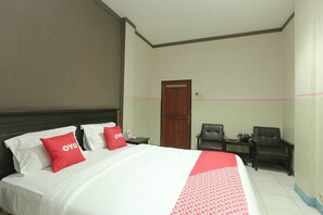 A hotel room with a large bed, two red pillows, a wooden headboard, a small table with a chair, and a door.
