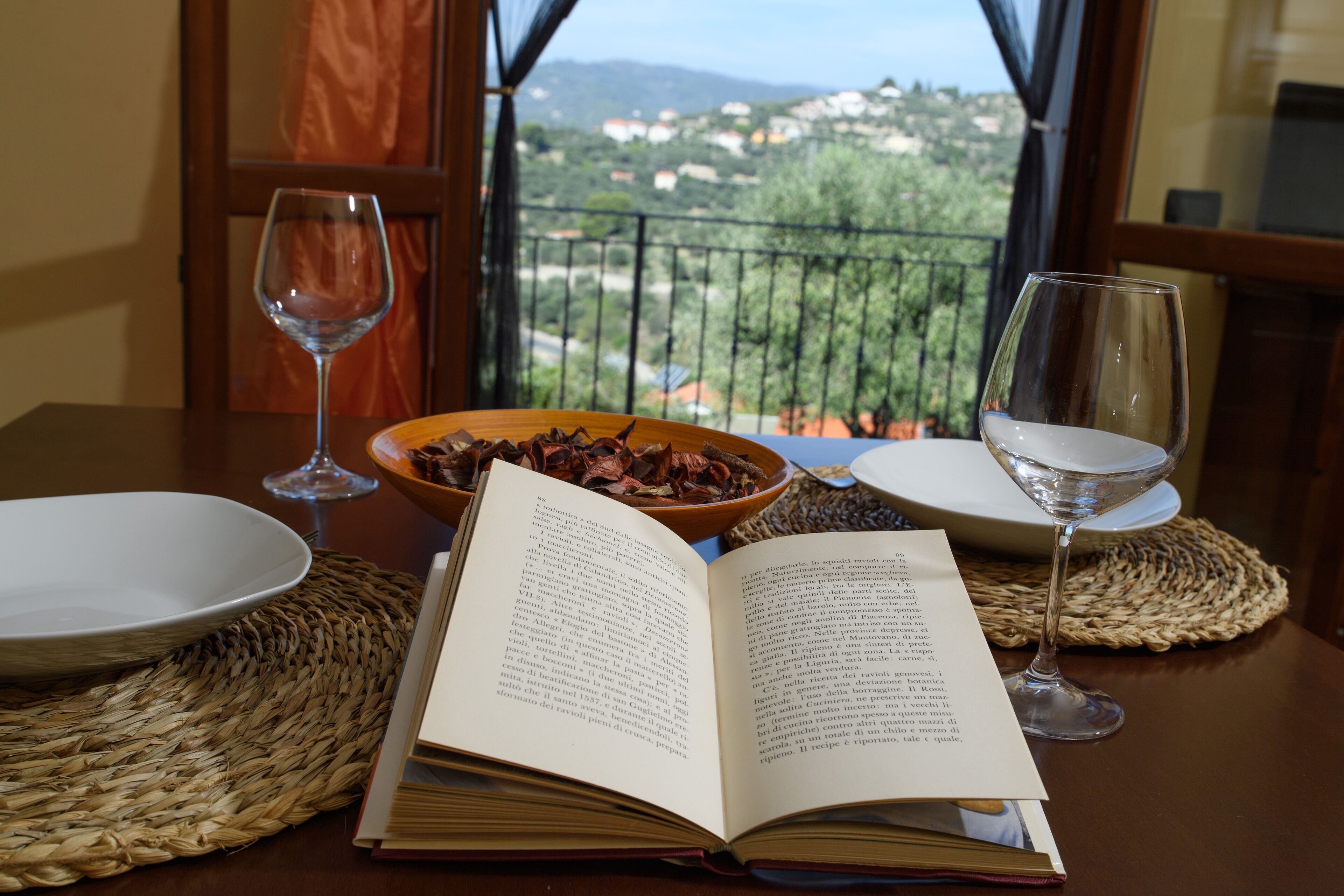 Una mesa de comedor con un libro, dos copas de vino y un bol de fruta seca.