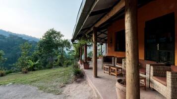 A covered outdoor seating area with wicker furniture and a stone fireplace, overlooking a lush landscape.
