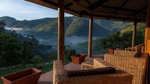 A covered outdoor seating area with wicker furniture and terracotta planters, overlooking a mountainous landscape with a misty valley. - Gorilla Valley Lodge (Bwindi)