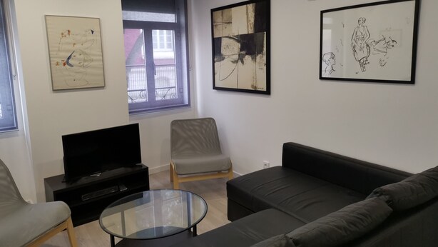 A modern living room with a black sofa, a glass coffee table, and two grey armchairs. There is a flat-screen TV on a black stand, and artwork is displayed on the walls.